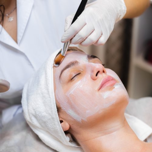 Beautician with a brush applies a white moisturizing mask to the face of a young girl client in a spa beauty salon Beautician with a brush applies a white moisturizing mask to the face of a young girl client in a spa beauty salon
